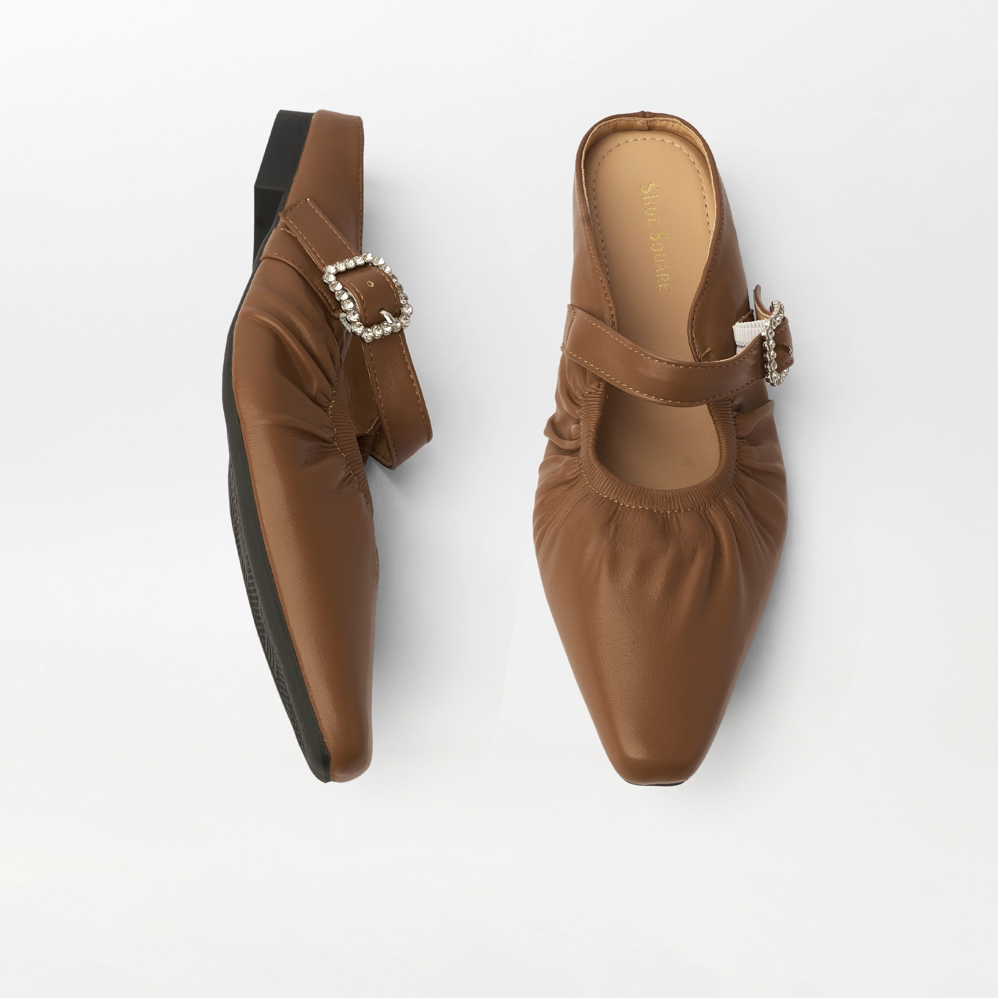 A pair of women's mule flats in smooth tan leather, shown from top and side angles on a clean white background. The shoes feature gathered detailing, a pointed toe, and a decorative buckle embellished with small rhinestones.
